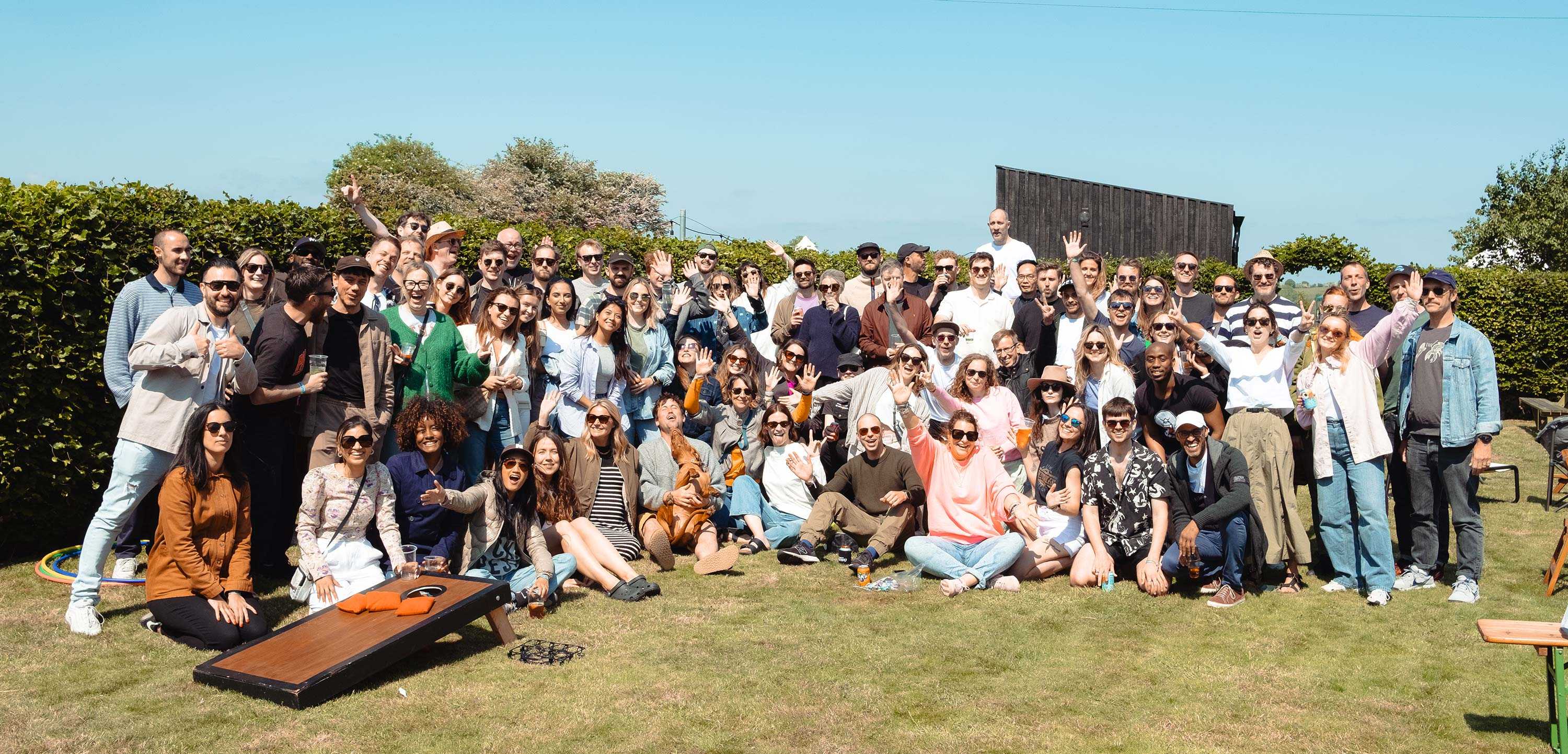 A group shot of employees at our annual social day - smiling, waving and cheering against a bucolic scene of green grass and blue sky