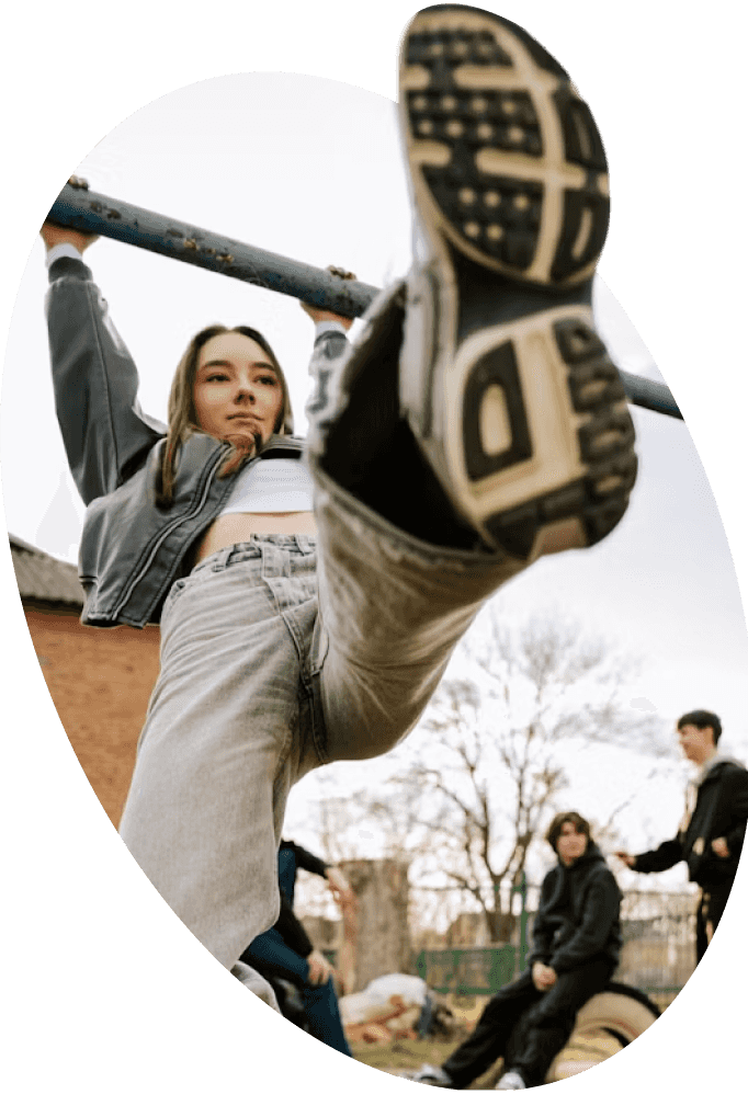 A young woman in jeans hangs from a metal exercise bar outdoors, lifting one leg high.
